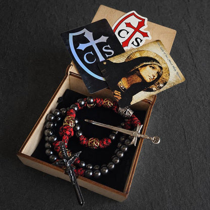 A boxed paracord rosary. The set includes a rosary with gunmetal beads, red and black paracord accents, pewter sword crucifix, a small sword, and devotional cards, all arranged inside an open wooden box.
