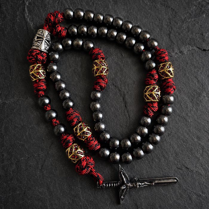 A paracord rosary with dark metallic beads, red and black braided cord, gold geometric spacer beads, a silver templar helmet bead, and a sword crucifix.