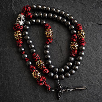 A paracord rosary with dark metallic beads, red and black braided cord, gold geometric spacer beads, a silver templar helmet bead, and a sword crucifix.