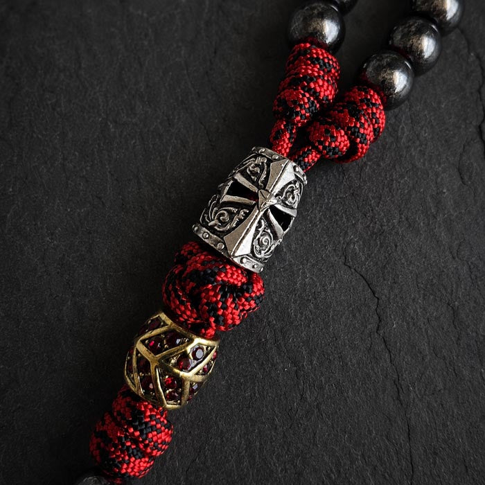 A close-up of a paracord rosary. The design features red and black braided cord, dark metallic round beads, a large silver-toned templar helmet centerpiece, and a smaller gold-toned geometric bead with rhinestone inlays.