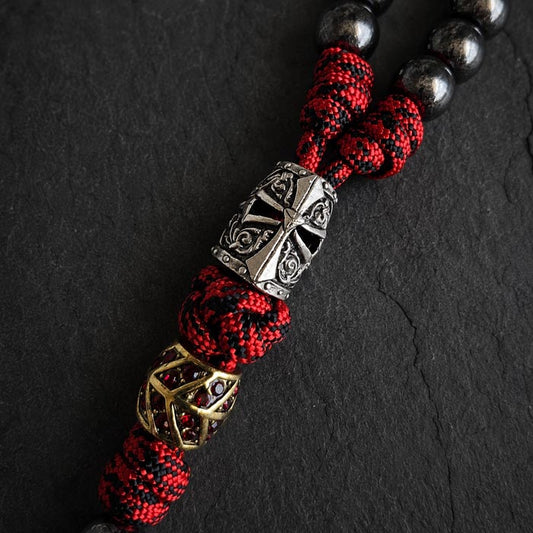 A close-up of a paracord rosary. The design features red and black braided cord, dark metallic round beads, a large silver-toned templar helmet centerpiece, and a smaller gold-toned geometric bead with rhinestone inlays.